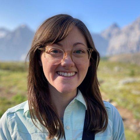 Head-and-shoulders portrait of an individual wearing eyeglasses and a light-colored button-down shirt, photographed outdoors. The background shows a grassy landscape with distant rocky mountains and a clear blue sky, softly blurred to emphasize the subject. Natural daylight illuminates the scene, with the individual centered in the frame.